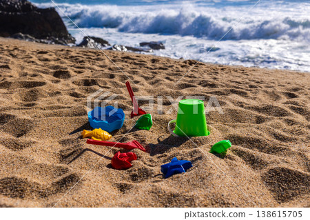 Plastic bucket with beach tools and molds placed on sand with sea and rocks in background. Outdoor beach photography. Summer vacation and seaside recreation concept. Wide angle shot Plastic bucket with beach tools and molds placed on sand with sea and rocks in background. Outdoor beach photography. Summer vacation and seaside recreation concept. Wide angle shot 138615705