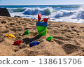 Plastic bucket with shovel and rake surrounded by sand molds on beach with waves in background. Outdoor beach photography. Summer vacation and seaside activity concept Eye level shot 138615706