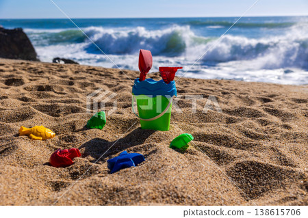 Plastic bucket with shovel and rake surrounded by sand molds on beach with waves in background. Outdoor beach photography. Summer vacation and seaside activity concept Eye level shot 138615706