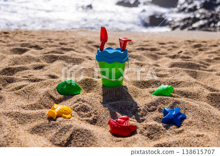 Children plastic bucket with shovel and rake on sandy beach near sea with toy molds around. Outdoor beach photography. Summer vacation and seaside play concept Eye level shot 138615707