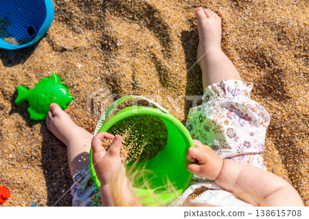 Toddler hands holding green bucket with sand pouring into it on beach. Outdoor beach photography. Childhood activity and summer vacation concept. Design for poster, banner, postcard. Top view shot 138615708