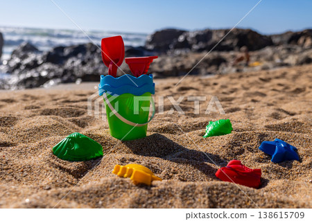 Plastic bucket with shovel and rake on sand with colorful toy molds on beach and sea background. Outdoor beach photography. Summer vacation and travel concept. Low angle shot 138615709