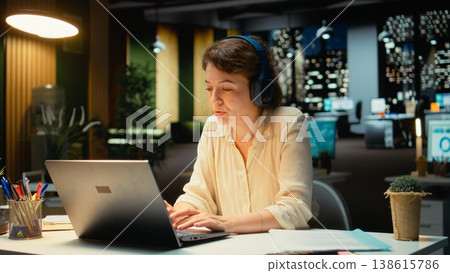 Businesswoman taking headphones out and stretching her back at desk, correcting her posture after working on tasks with ambience music. Woman with muscle stiffness and tension. Camera A. Businesswoman taking headphones out and stretching her back at desk, correcting her posture after working on tasks with ambience music. Woman with muscle stiffness and tension. Camera A. 138615786