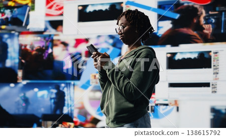 Happy woman uses smartphone to do texting with friends, discussing movies, standing in front of vibrant video wall collage. Teenager chatting with other social media users using mobile phone, camera B Happy woman uses smartphone to do texting with friends, discussing movies, standing in front of vibrant video wall collage. Teenager chatting with other social media users using mobile phone, camera B 138615792