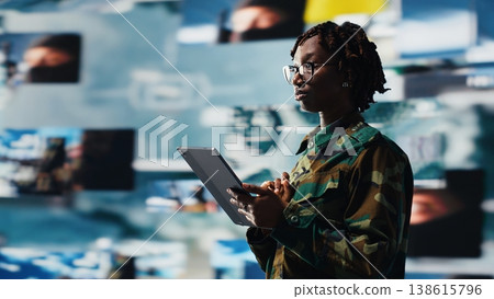 Soldier providing combat support, looking at reconnaissance feeds on videowall with tablet app. Intelligence agency professional uses battlefield communication network on device, camera B 138615796