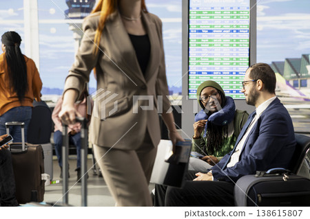Diverse passengers socializing while they wait for boarding flight, sitting with luggage in busy airport terminal lounge. Suitcases in departure waiting area near gate, board plane for journey. 138615807