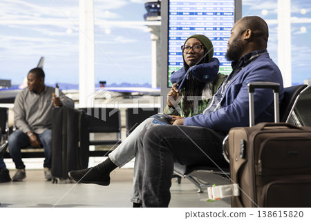 Airport terminal travelers, black people socializing, international travel vacation, waiting for departure in lounge area. Couple with luggage relaxing on bench before boarding their plane. Airport terminal travelers, black people socializing, international travel vacation, waiting for departure in lounge area. Couple with luggage relaxing on bench before boarding their plane. 138615820