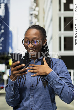 Close up of jolly woman laughing at funny online clips on smartphone, walking outside. Smiling African american young girl on city street amused by hilarious memes on mobile phone Close up of jolly woman laughing at funny online clips on smartphone, walking outside. Smiling African american young girl on city street amused by hilarious memes on mobile phone 138615824