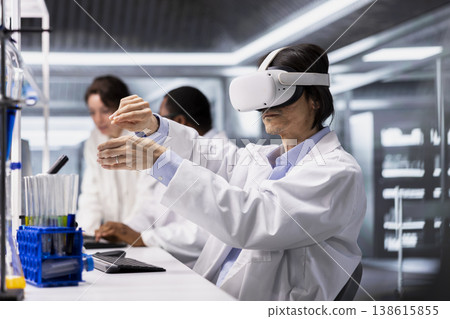 Lab employee wearing VR headset gear using chemical fluids for experiments. Chemist uses virtual reality tech to simulate using indicator solution laboratory glassware filled at workstation Lab employee wearing VR headset gear using chemical fluids for experiments. Chemist uses virtual reality tech to simulate using indicator solution laboratory glassware filled at workstation 138615855