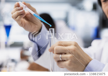 Close up of chemical engineer using pipette to transfer liquid into test tube containing blue solution at laboratory bench. Research lab specialist does pipetting procedure with chemical sample Close up of chemical engineer using pipette to transfer liquid into test tube containing blue solution at laboratory bench. Research lab specialist does pipetting procedure with chemical sample 138615875
