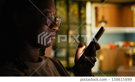 African American young woman relaxing alone and checking apps, scrolling social media on phone at home. Comfortable domestic life highlighting calm cozy lifestyle, modern everyday tranquility. African American young woman relaxing alone and checking apps, scrolling social media on phone at home. Comfortable domestic life highlighting calm cozy lifestyle, modern everyday tranquility. 138615899
