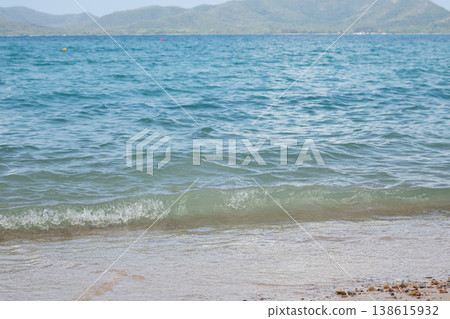 A soft green-blue wave curls along a sandy shore with pebbles, while distant hills fade into the horizon, offering a serene seaside atmosphere perfect for leisure and coastal travel themes. 138615932