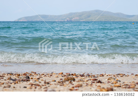 Clear blue ocean waves softly break on a sandy shore scattered with pebbles, with a calm island landscape in the distance under bright daylight, creating a peaceful coastal scene. Clear blue ocean waves softly break on a sandy shore scattered with pebbles, with a calm island landscape in the distance under bright daylight, creating a peaceful coastal scene. 138616082