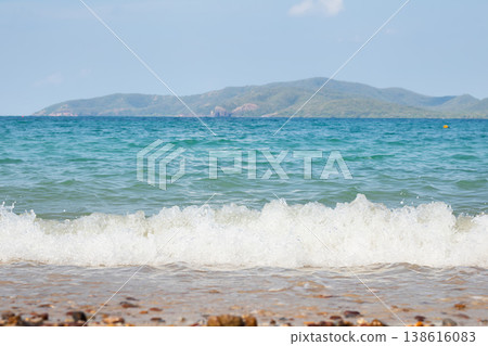 Vivid turquoise sea wave splashes onto a sandy pebble beach, with a lush island stretching across the horizon under a bright blue sky, capturing a refreshing tropical coastal atmosphere. 138616083