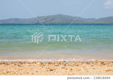 Soft emerald-green seawater gently approaches a sandy beach covered with small stones, while a hazy mountain island stretches across the horizon beneath a clear and peaceful sky. 138616084