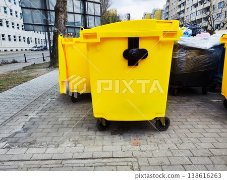 Yellow garbage bins with wheels are placed on a paved sidewalk in an urban setting, a communal waste collection area 138616263