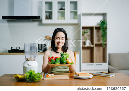 Happy Asian woman holding a plate of fresh vegetables and smiling in a modern kitchen. Concept of healthy lifestyle, clean eating, vegan diet, and organic nutrition 138620867