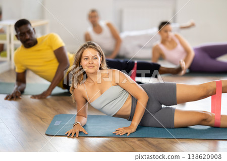 Girl with friends perform exercises lying on side and narrow elastic band on shins Girl with friends perform exercises lying on side and narrow elastic band on shins 138620908