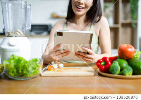 Smiling Asian woman using tablet surrounded by fresh vegetables in kitchen. Concept of healthy lifestyle, cooking, blogging, and digital wellness Smiling Asian woman using tablet surrounded by fresh vegetables in kitchen. Concept of healthy lifestyle, cooking, blogging, and digital wellness 138620976
