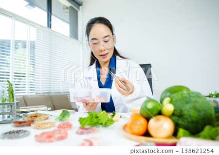Female scientist in lab coat and goggles examining raw meat with tweezers in food lab. Nutrition, protein, food safety, and research concept. Female scientist in lab coat and goggles examining raw meat with tweezers in food lab. Nutrition, protein, food safety, and research concept. 138621254