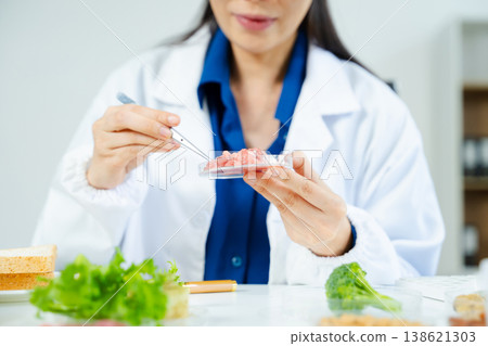 Female scientist in lab coat and goggles examining raw meat with tweezers in food lab. Nutrition, protein, food safety, and research concept. 138621303