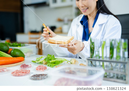 Female nutritionist in lab coat analyzing bread with vegetables, meat, nuts, and grains. Food science, healthy diet, and nutrition research concept. Female nutritionist in lab coat analyzing bread with vegetables, meat, nuts, and grains. Food science, healthy diet, and nutrition research concept. 138621324