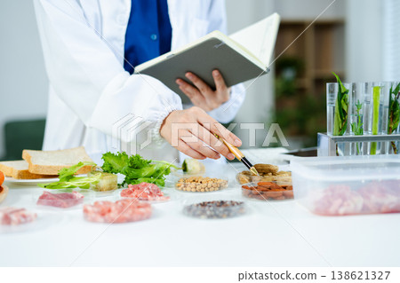 Female nutritionist in lab coat analyzing food and writing in notebook. Healthy eating, organic testing, and nutrition science in a clean lab. Female nutritionist in lab coat analyzing food and writing in notebook. Healthy eating, organic testing, and nutrition science in a clean lab. 138621327