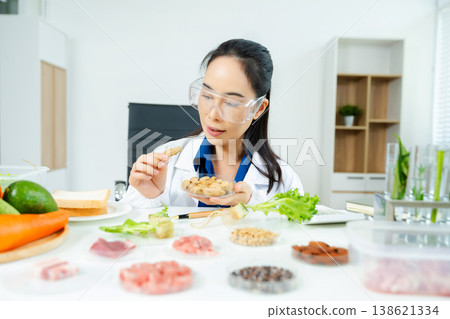Female nutritionist in lab coat and goggles inspecting green plant in test tube. Food research, health, nutrition science, and biotech lab concept. Real people in clinical setting. Female nutritionist in lab coat and goggles inspecting green plant in test tube. Food research, health, nutrition science, and biotech lab concept. Real people in clinical setting. 138621334