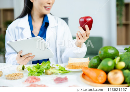 Smiling female nutritionist holding apple and digital tablet while analyzing healthy foods. Food science, diet planning, and health research concept. Real people in lab setting. Smiling female nutritionist holding apple and digital tablet while analyzing healthy foods. Food science, diet planning, and health research concept. Real people in lab setting. 138621367