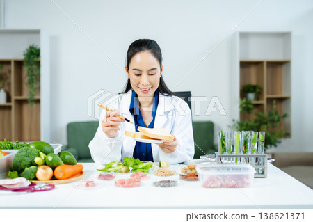 Female nutritionist in lab coat analyzing bread with vegetables, meat, nuts, and grains. Food science, healthy diet, and nutrition research concept. 138621371