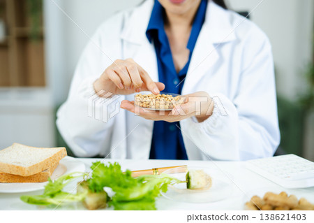 Nutrition expert in lab coat presenting soybeans and almonds in petri dishes. Healthy food, protein, diet, and food science concept. Clinical setting Nutrition expert in lab coat presenting soybeans and almonds in petri dishes. Healthy food, protein, diet, and food science concept. Clinical setting 138621403