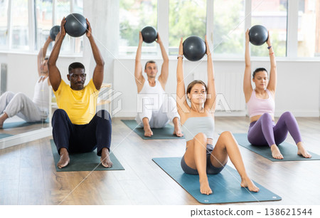 Pilates class in multi-ethnic group - men and women hold the ball above their heads and do bends Pilates class in multi-ethnic group - men and women hold the ball above their heads and do bends 138621544