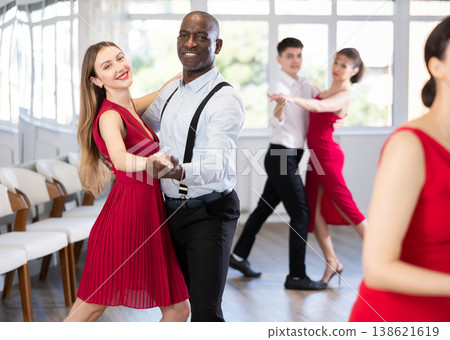Positive woman in red dress and African american man practicing waltz dance moves in pair during group party in dancing studio 138621619