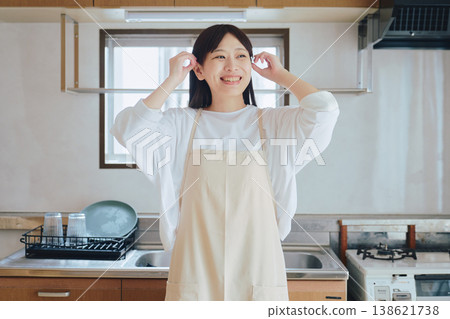 一位身穿圍裙的女子在廚房裡微笑;一幅家務和烹飪的畫面;一幅積極向上的生活方式場景。 一位身穿圍裙的女子在廚房裡微笑;一幅家務和烹飪的畫面;一幅積極向上的生活方式場景。 138621738