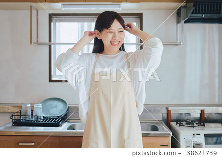 一位身穿圍裙的女子在廚房裡微笑;一幅家務和烹飪的畫面;一幅積極向上的生活方式場景。 一位身穿圍裙的女子在廚房裡微笑;一幅家務和烹飪的畫面;一幅積極向上的生活方式場景。 138621739