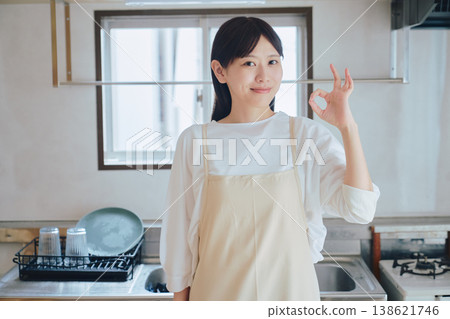 一位身穿圍裙的女子在廚房裡微笑;一幅家務和烹飪的畫面;一幅積極向上的生活方式場景。 一位身穿圍裙的女子在廚房裡微笑;一幅家務和烹飪的畫面;一幅積極向上的生活方式場景。 138621746