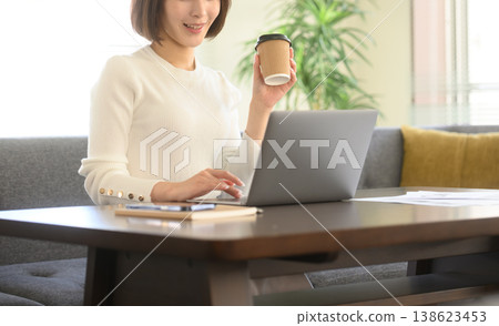 A woman is at home, using her laptop and drinking coffee. A woman is at home, using her laptop and drinking coffee. 138623453