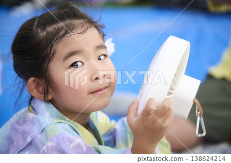 A girl in a yukata holding a handheld fan and looking at the camera. 138624214