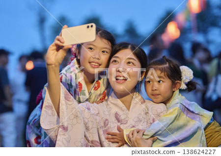 A mother and her daughters, both dressed in yukata, taking photos with their smartphones at a summer festival. A mother and her daughters, both dressed in yukata, taking photos with their smartphones at a summer festival. 138624227