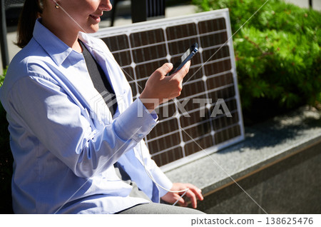Happy woman using smartphone connected to solar panel. Cropped view. Integration of sustainable renewable energy into everyday life, demonstrating practical use of solar power for charging devices. Happy woman using smartphone connected to solar panel. Cropped view. Integration of sustainable renewable energy into everyday life, demonstrating practical use of solar power for charging devices. 138625476