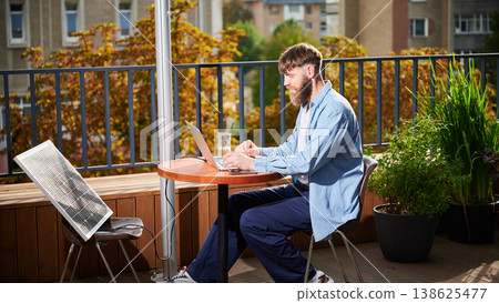 Man works on laptop at outdoor round table, with glass of iced coffee. Solar panel nearby, emphasizing sustainable, modern eco-friendly workspace that combines technology and sustainability. 138625477