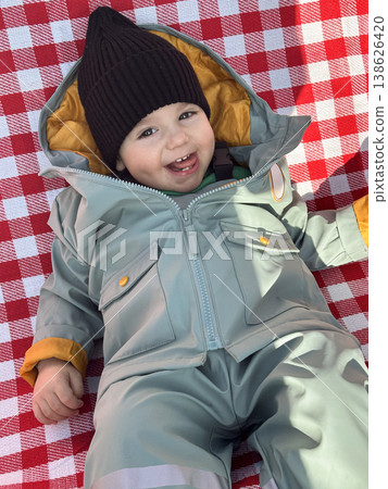 Smiling toddler in winter snowsuit on red checkered blanket Smiling toddler in winter snowsuit on red checkered blanket 138626420