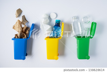 Sorting and recycling waste, three toy recycling bins for glass, paper, and plastic on white background top view. Color coded containers promoting eco friendly waste sorting and sustainable living. 138628614