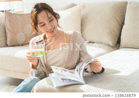 一位女士坐在沙發上，一邊看雜誌，一邊喝檸檬水和排毒水。 138628879