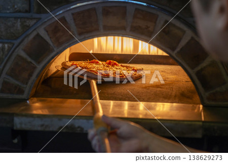 A scene of baking pizza in a stone oven; the pizza is being baked using a pizza peel in an authentic pizza oven with flames burning. A scene of baking pizza in a stone oven; the pizza is being baked using a pizza peel in an authentic pizza oven with flames burning. 138629273