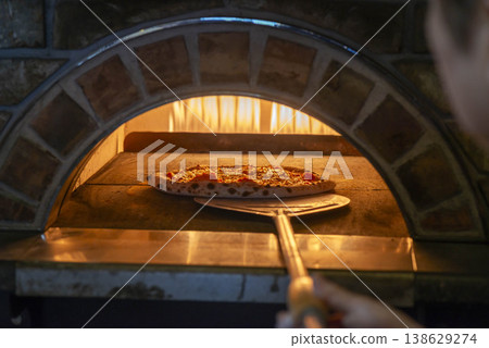 A scene of baking pizza in a stone oven; the pizza is being baked using a pizza peel in an authentic pizza oven with flames burning. A scene of baking pizza in a stone oven; the pizza is being baked using a pizza peel in an authentic pizza oven with flames burning. 138629274