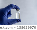 A hand wearing a blue nitrile glove holding a small clear glass vial with a metal cap 138629970