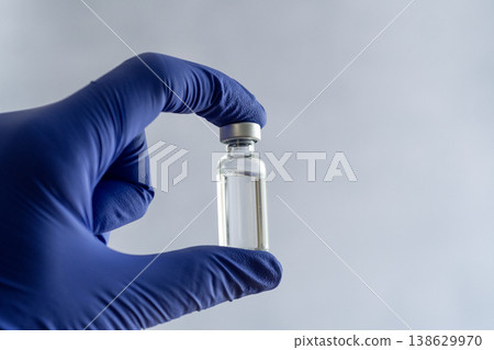 A hand wearing a blue nitrile glove holding a small clear glass vial with a metal cap A hand wearing a blue nitrile glove holding a small clear glass vial with a metal cap 138629970