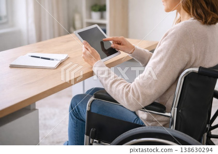 Woman in wheelchair using digital tablet at desk in bright modern home Woman in wheelchair using digital tablet at desk in bright modern home 138630294