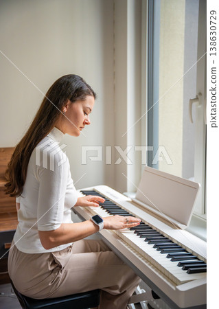 Young woman playing on white piano synthesizer at home. Concept of home lesson of music: learning music enhances motor skills, hearing, and intelligence, fostering creativity Young woman playing on white piano synthesizer at home. Concept of home lesson of music: learning music enhances motor skills, hearing, and intelligence, fostering creativity 138630729